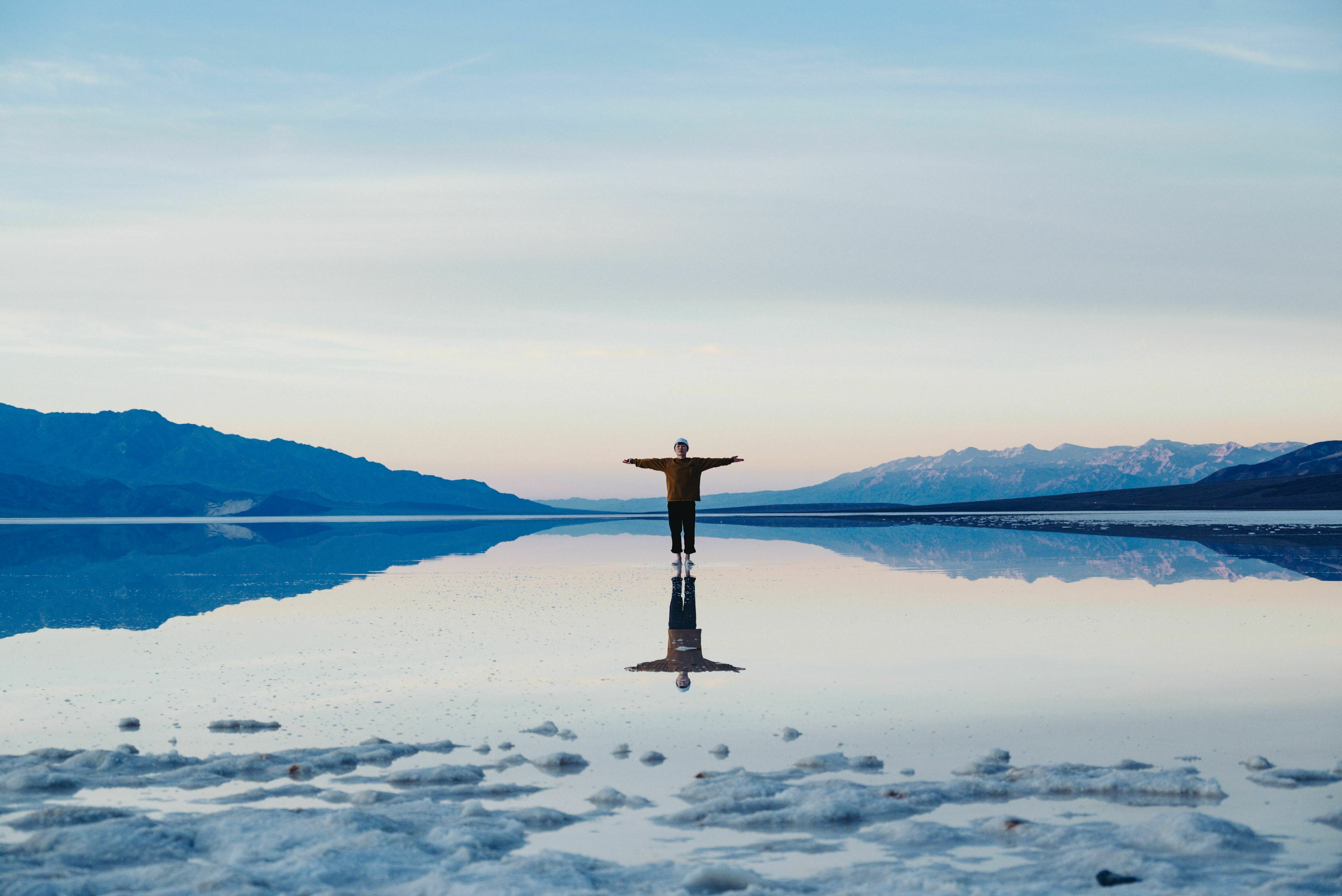 a wide panorama landscape with a sea and mountains in the background, in the center of the picture a person is standing with their arms spread out