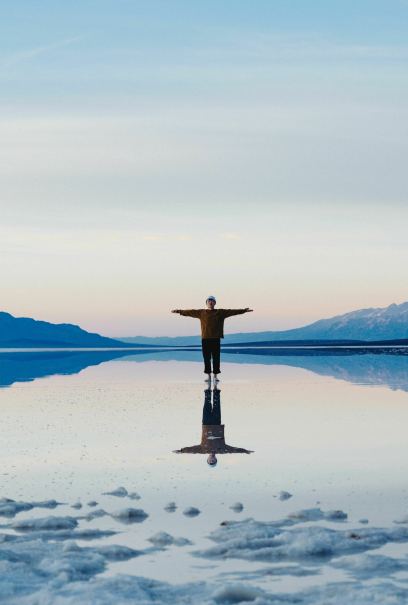 A person stands with arms outstretched, reflected in a calm body of water. Surrounding mountains and a pastel sky.